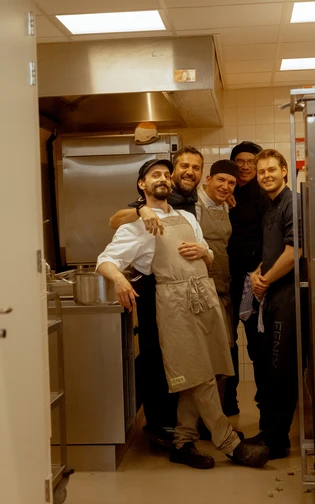 Five smiling chefs pose together in a commercial kitchen. They wear aprons and chef hats