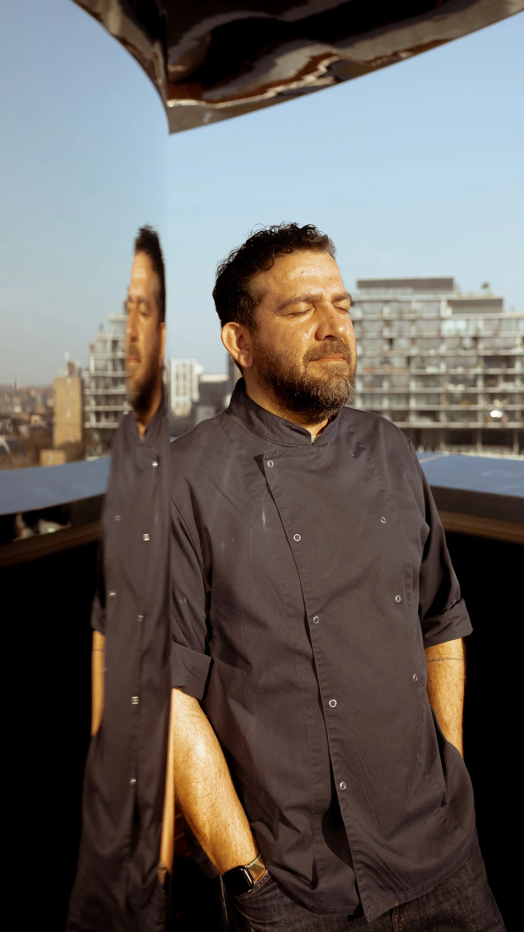 A man in a navy chef's coat stands with eyes closed, reflecting against a mirrored surface.