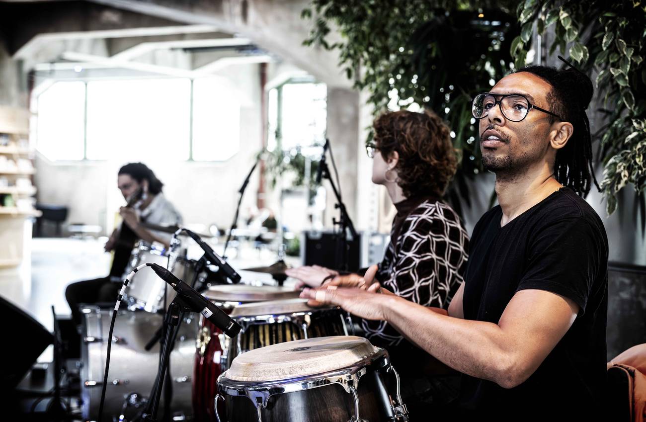 Three musicians performing in a bright room with plants. Focus on a person playing congas passionate