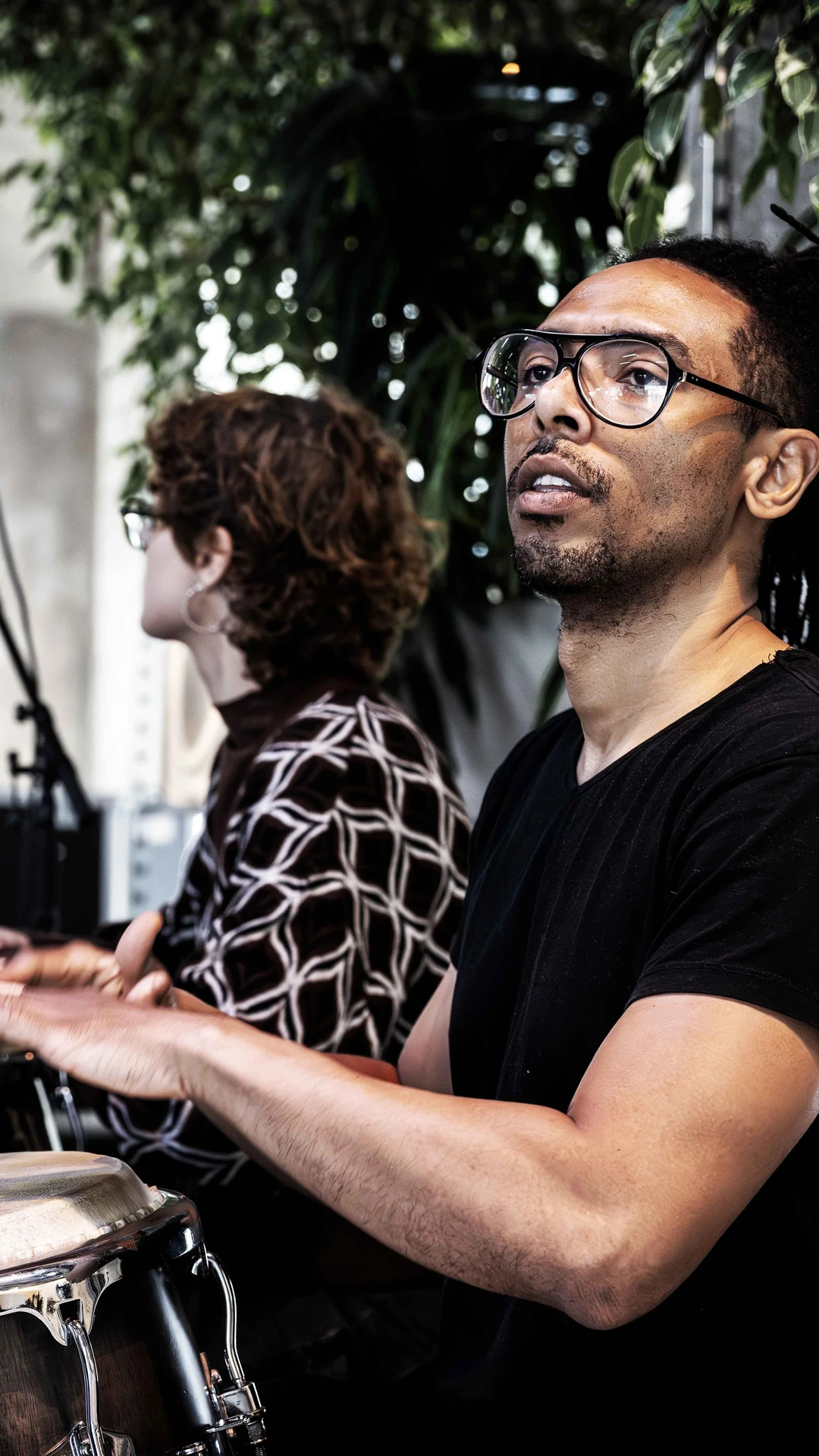 Three musicians performing in a bright room with plants. Focus on a person playing congas passionate