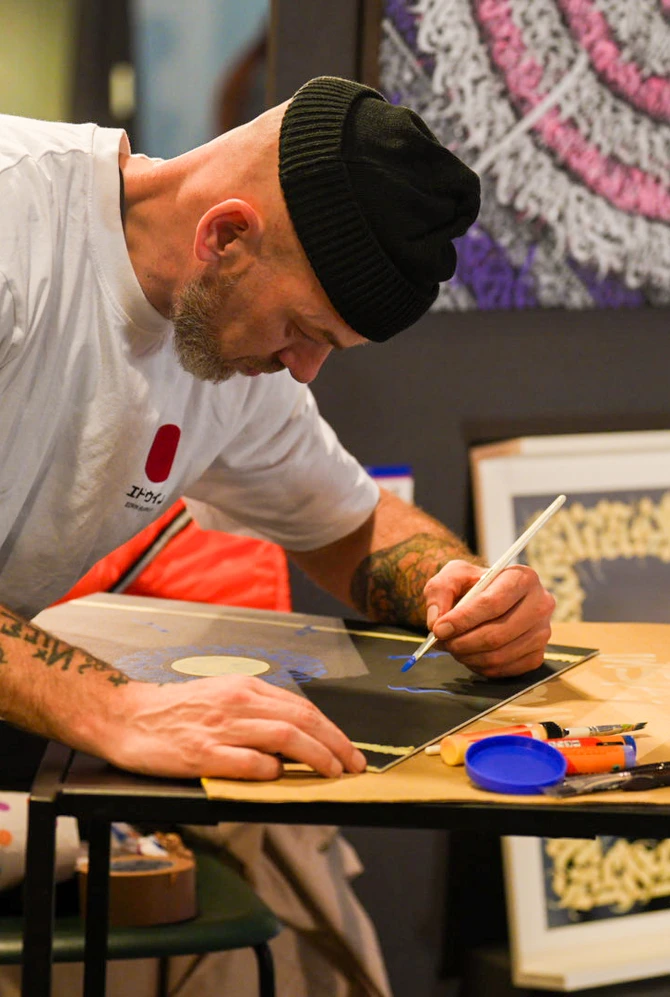 A man in a beanie and tattooed arms paints intently on a small table.
