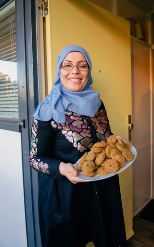A smiling woman in a light blue hijab and patterned dress holds a plate of cookies.