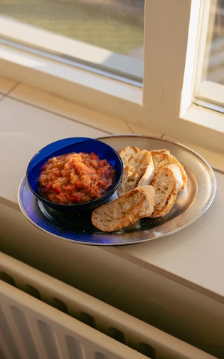 A blue bowl filled with chunky red salsa is on a round metal tray alongside slices of rustic bread