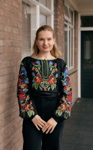 A woman stands smiling in a hallway. She is wearing a black blouse with vibrant floral embroidery.