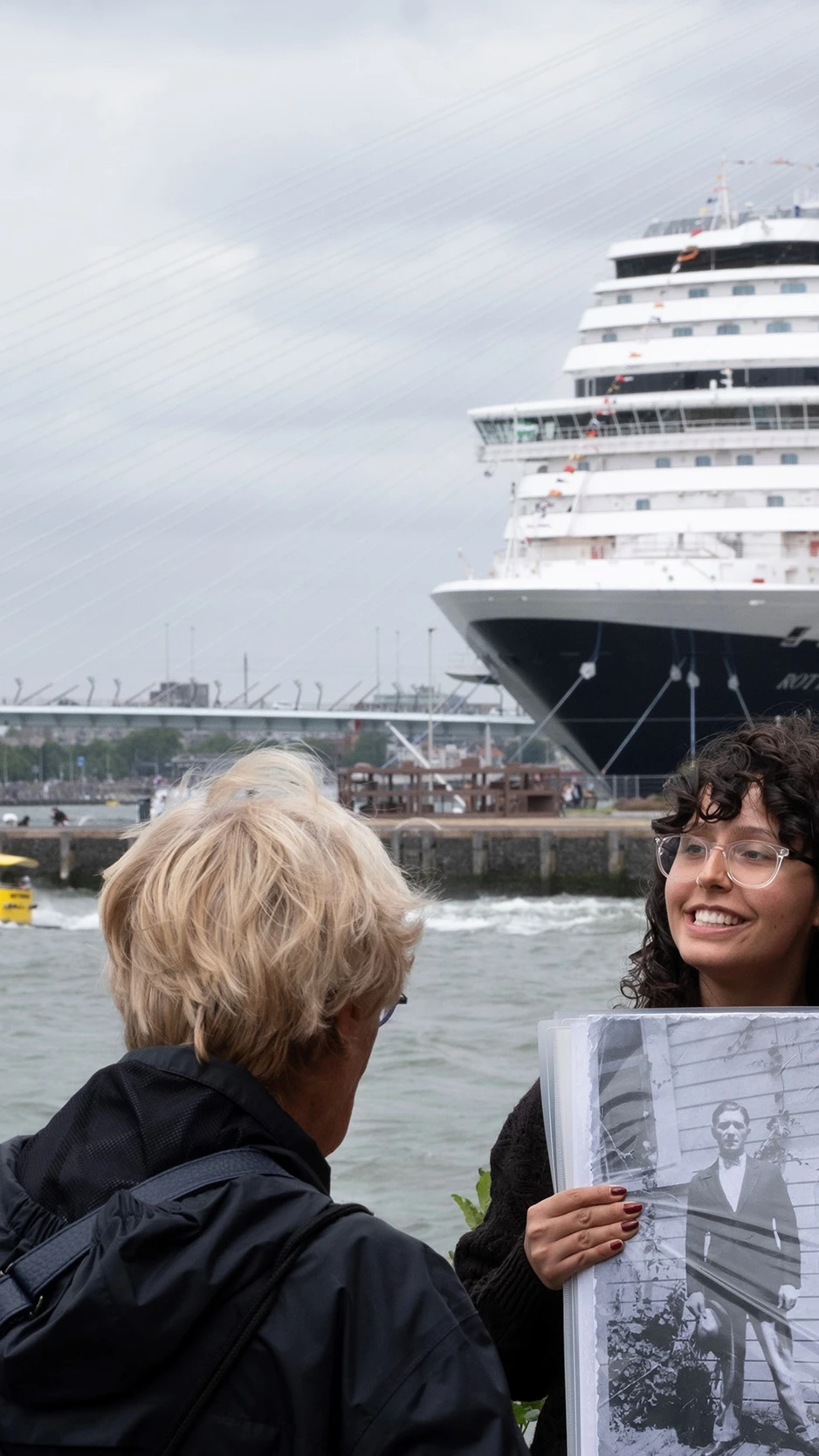 Een gids houdt buiten aan de Rotterdamse kade een zwart-witfoto omhoog voor een kleine groep luister