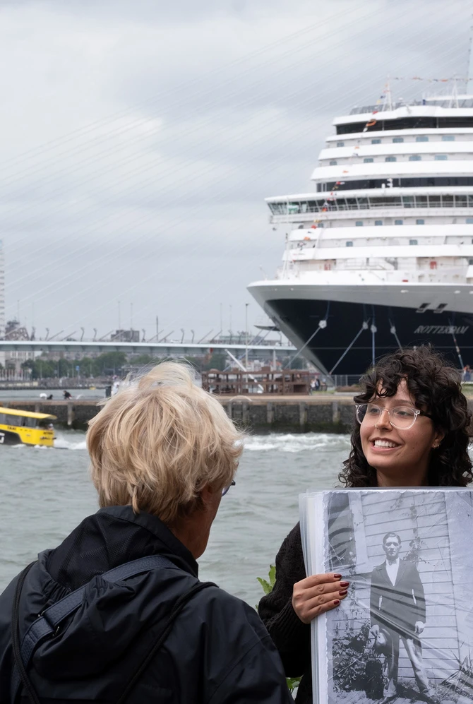 Een gids houdt buiten aan de Rotterdamse kade een zwart-witfoto omhoog voor een kleine groep luister