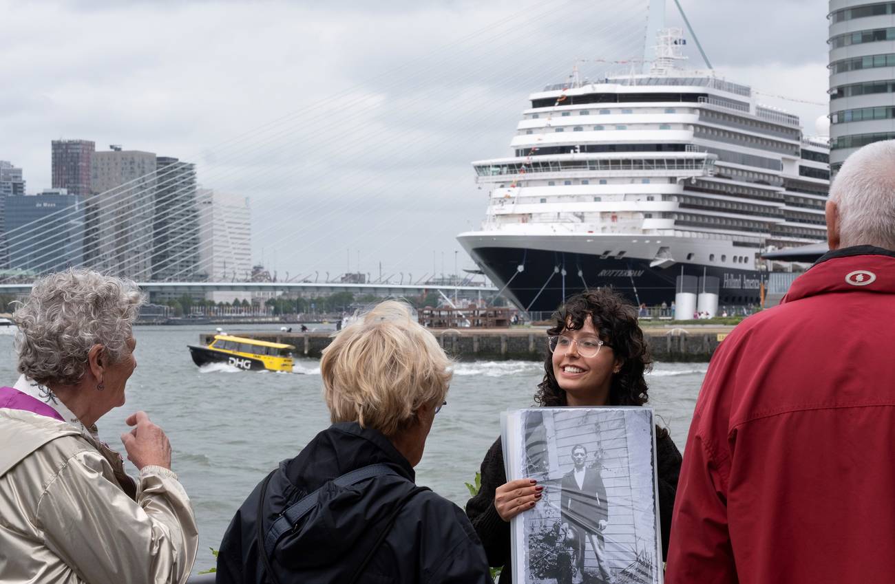 Een gids houdt buiten aan de Rotterdamse kade een zwart-witfoto omhoog voor een kleine groep luister