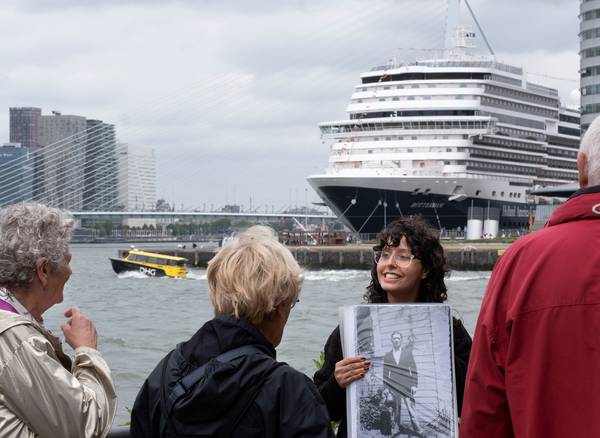 Een gids houdt buiten aan de Rotterdamse kade een zwart-witfoto omhoog voor een kleine groep luister