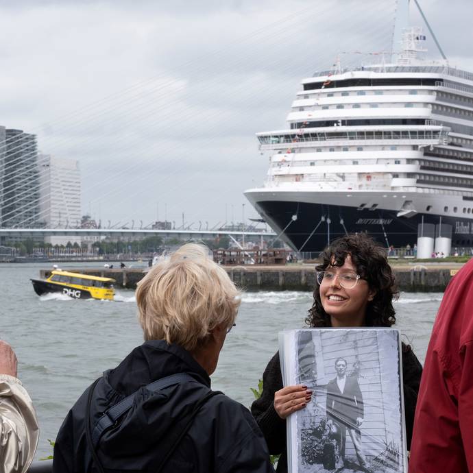Een gids houdt buiten aan de Rotterdamse kade een zwart-witfoto omhoog voor een kleine groep luister