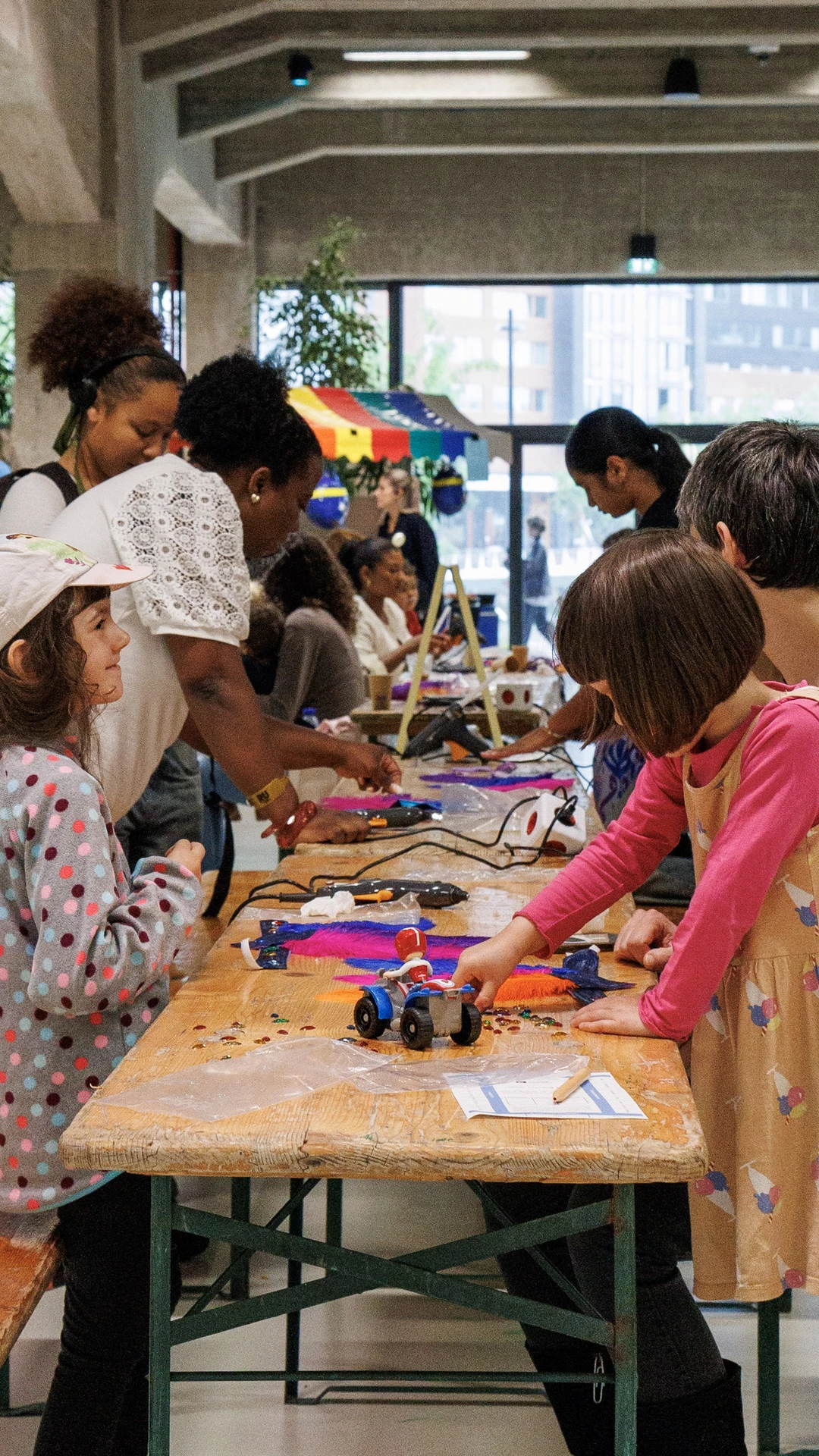 Children and adults engage in arts and crafts at a long wooden table indoors.