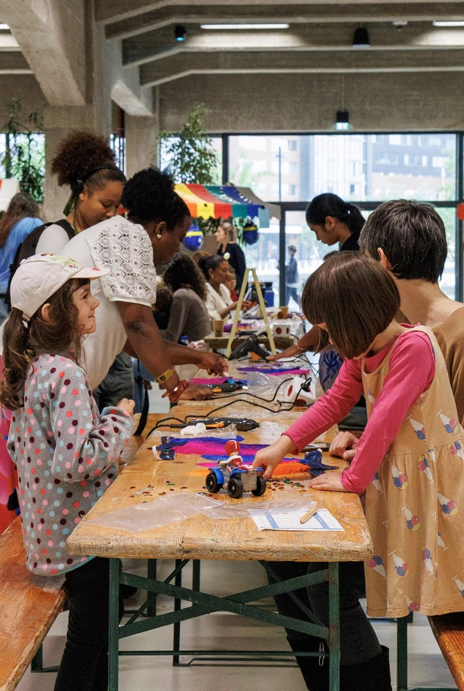 Children and adults engage in arts and crafts at a long wooden table indoors.