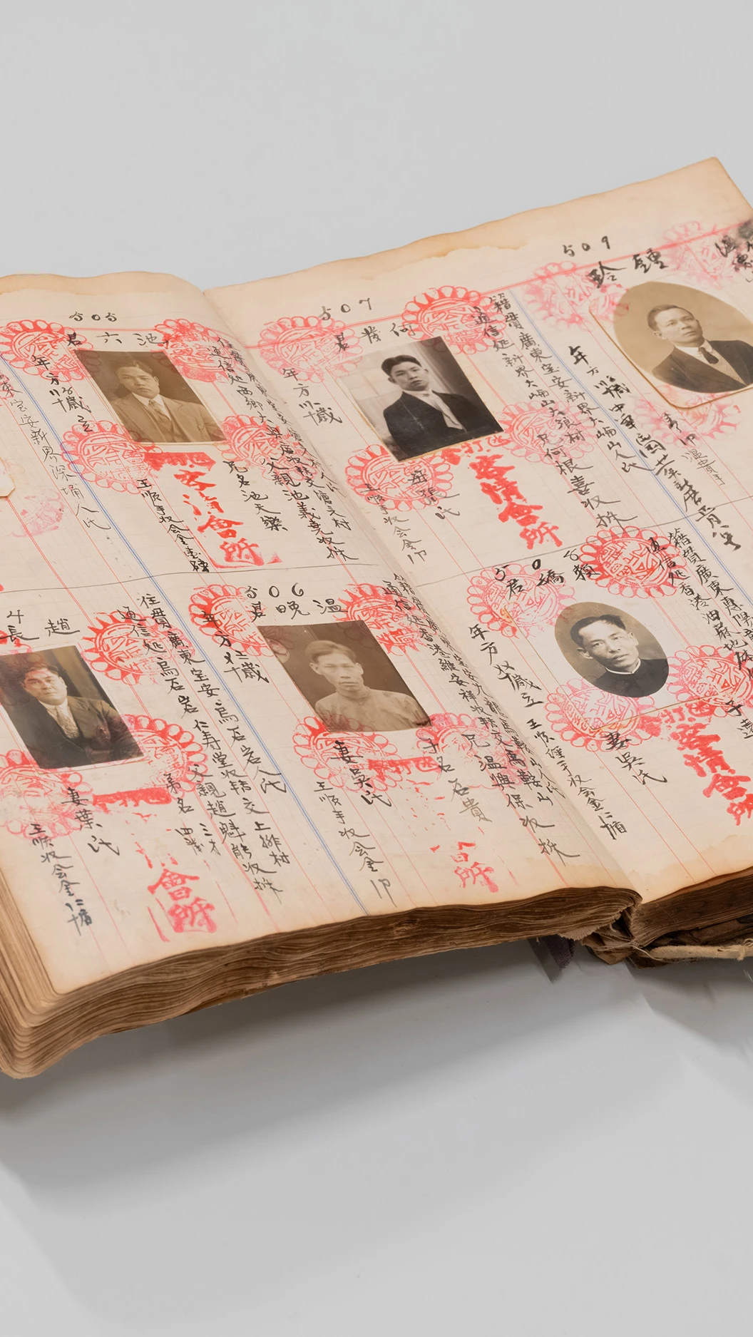 een oud boek met pasfoto's, Chinees schrift en rode stempels ligt open op een grijze ondergrond