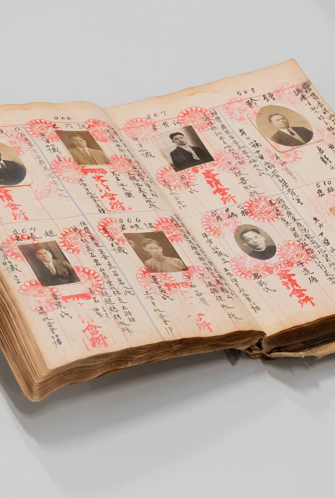 een oud boek met pasfoto's, Chinees schrift en rode stempels ligt open op een grijze ondergrond