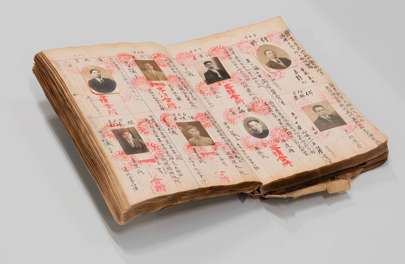een oud boek met pasfoto's, Chinees schrift en rode stempels ligt open op een grijze ondergrond