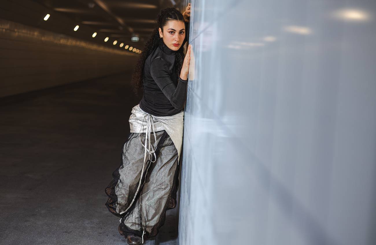 Woman in a black top and layered skirt leans against a shiny wall in an underground tunnel.