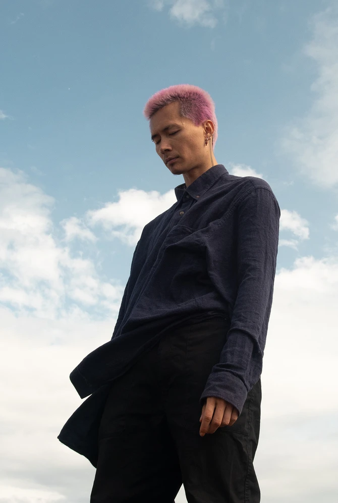 A person with short pink hair stands against a cloudy blue sky, wearing a dark shirt and trousers.