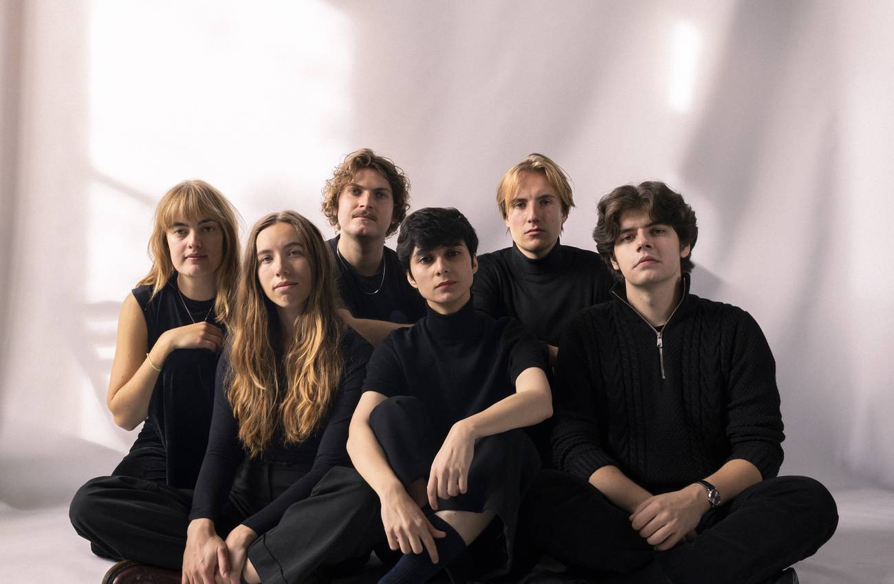 Six members of Mahala dressed in black sit together against a backdrop.