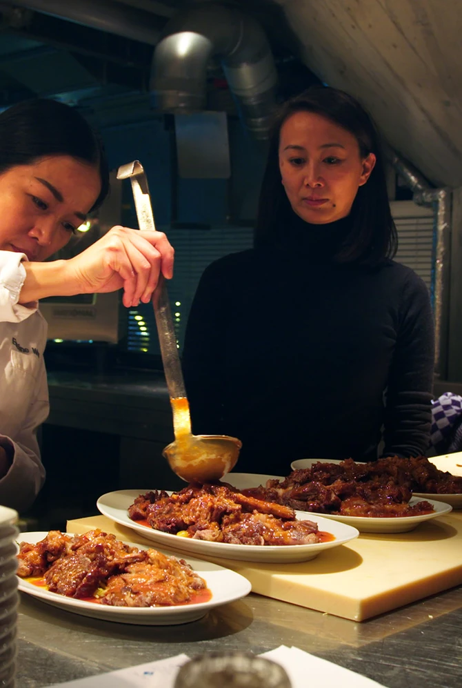 A chef in a white coat carefully ladles sauce over plated meat dishes in a restaurant kitchen.