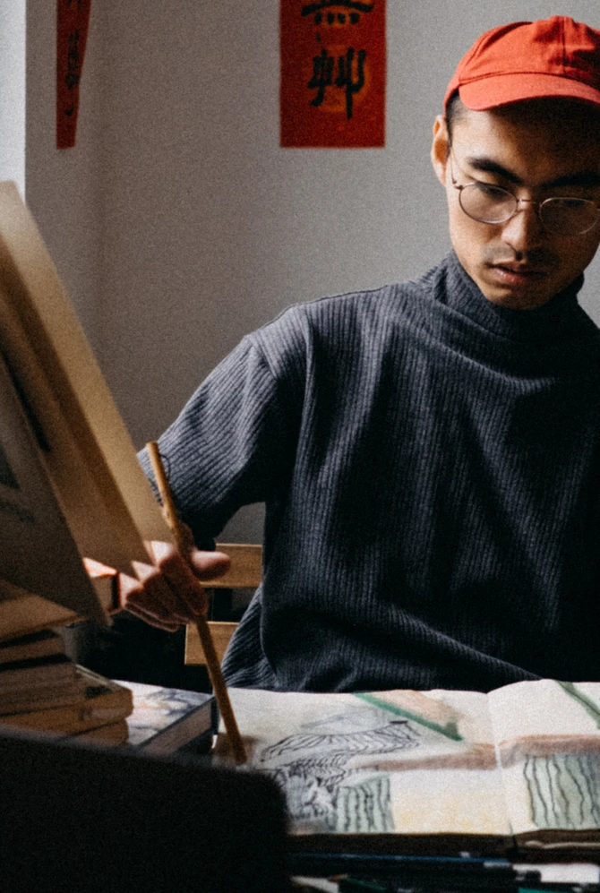 An illustrator works on a drawing at a table beneath Chinese calligraphy.