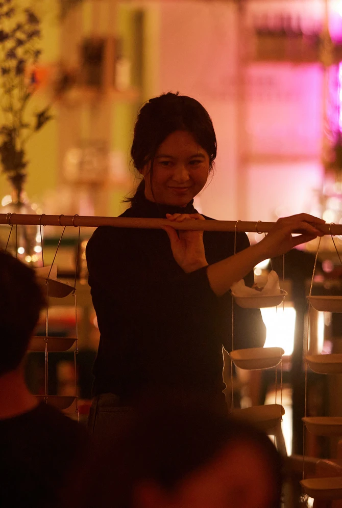 A woman in a dimly-lit, cozy restaurant balances a wooden rod hung with small bowls on strings