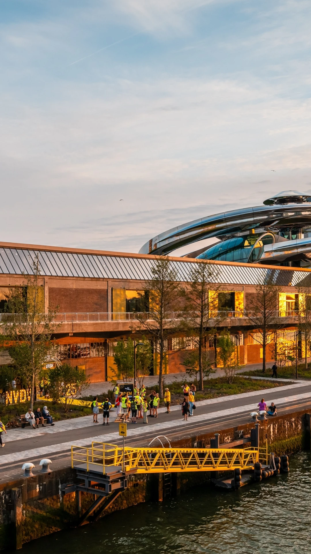 Sunset view of a modern riverside building with a futuristic rooftop structure in Rotterdam. People