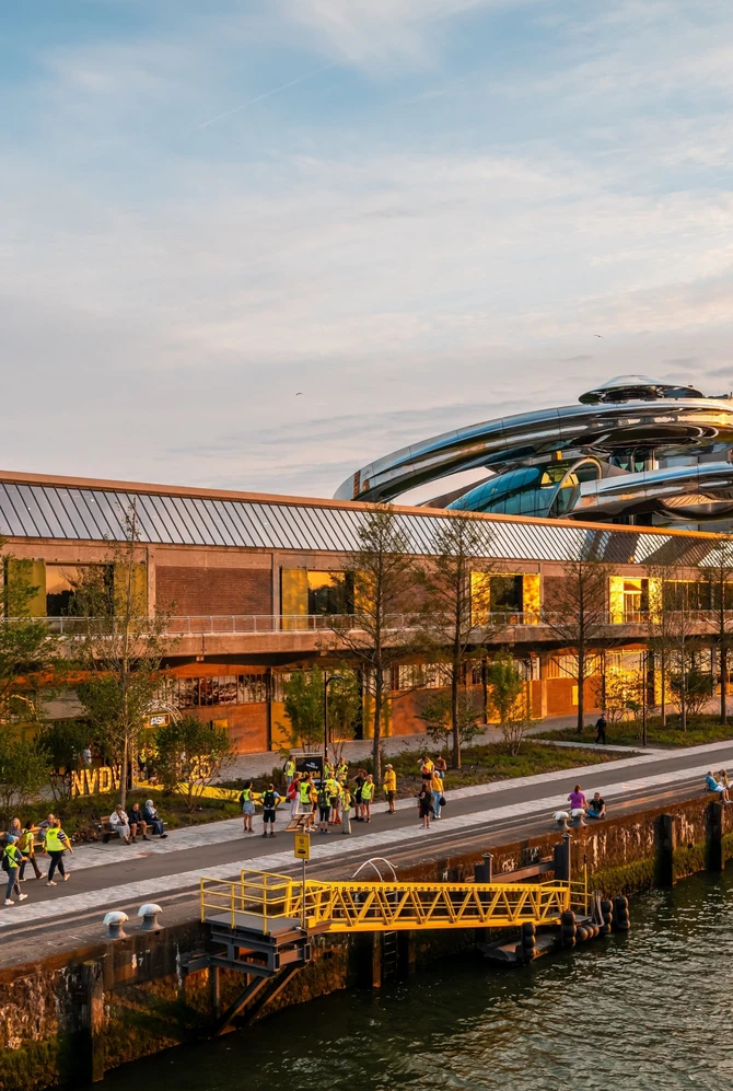 Sunset view of a modern riverside building with a futuristic rooftop structure in Rotterdam. People