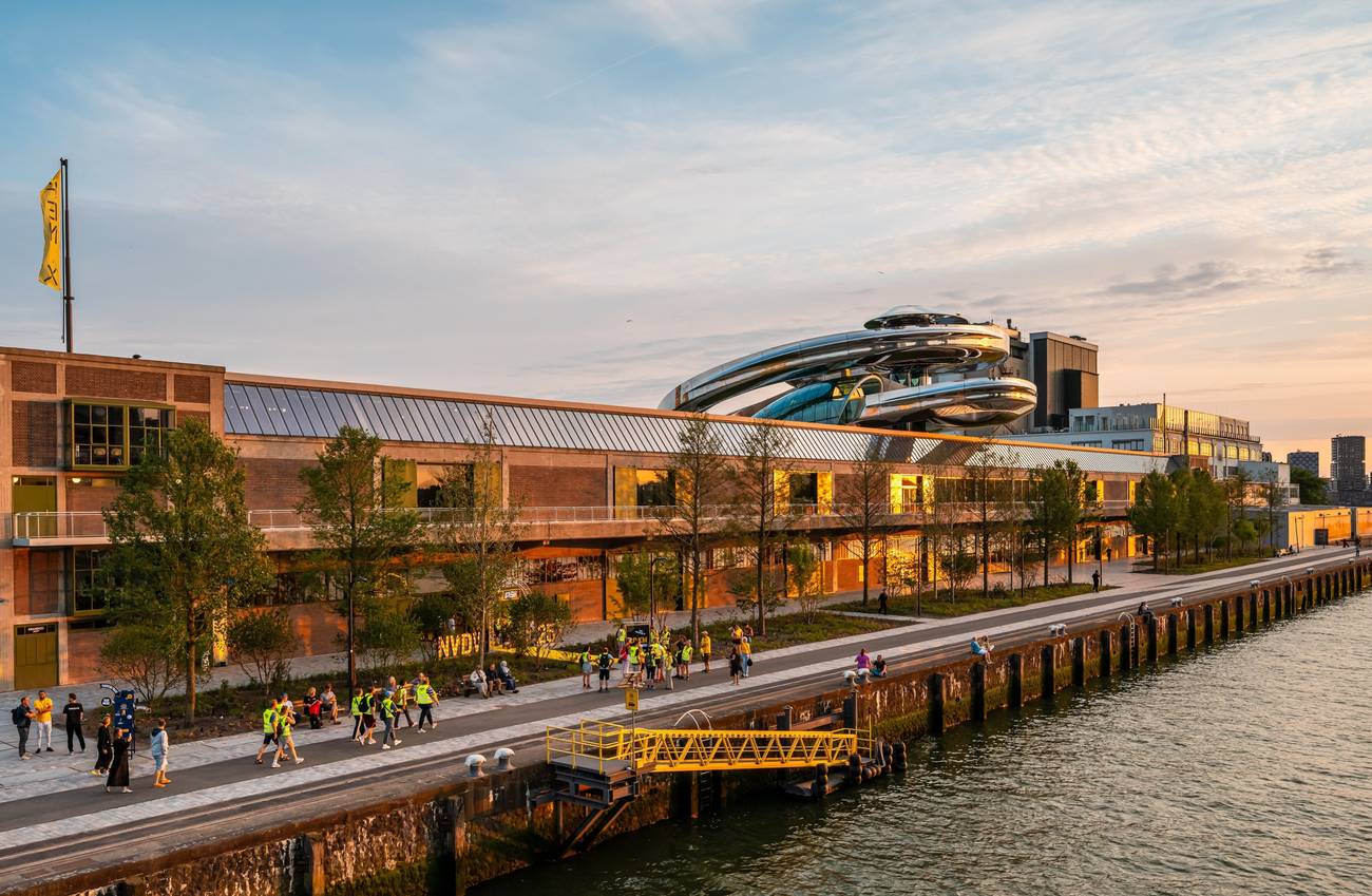 Sunset view of a modern riverside building with a futuristic rooftop structure in Rotterdam. People