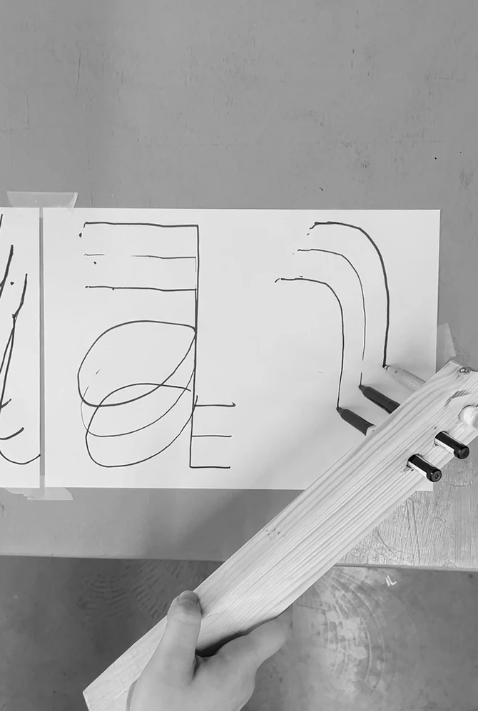 A hand-held wooden drawing tool creates abstract black line patterns on paper.
