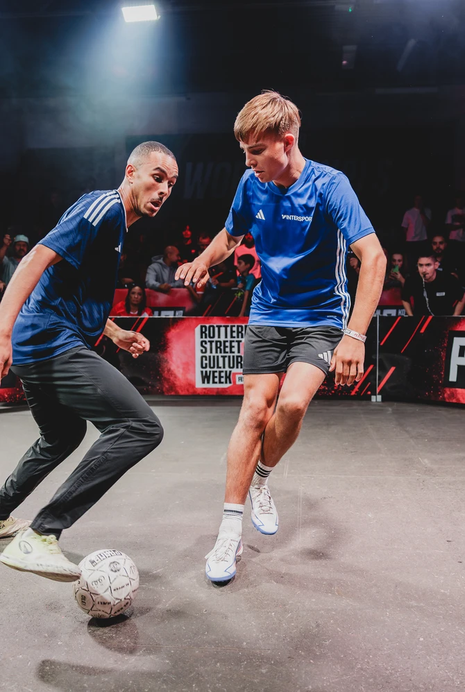 Two street football players battle for the ball in a panna arena as spectators watch.