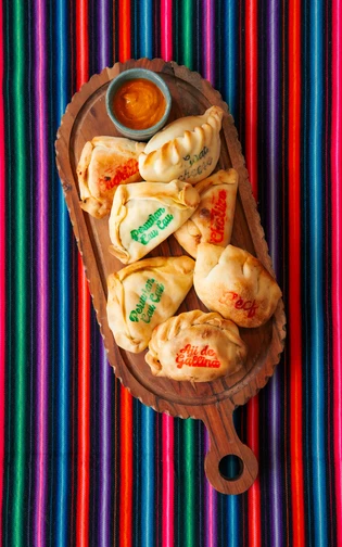 Houten schaal met verschillende empanada’s met namen op de korst en een kommetje saus ernaast.