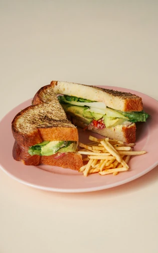Broodje op een roze bord, doormidden gesneden, met avocado en chips.