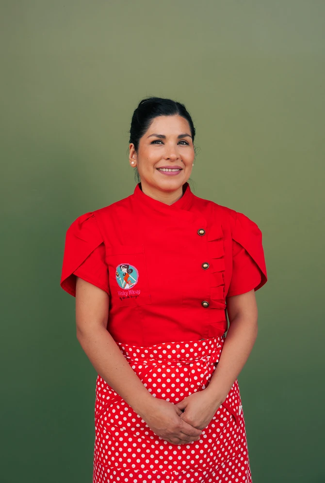 A woman wearing a red chef's jacket and a polka dot apron, smiling against a green background.