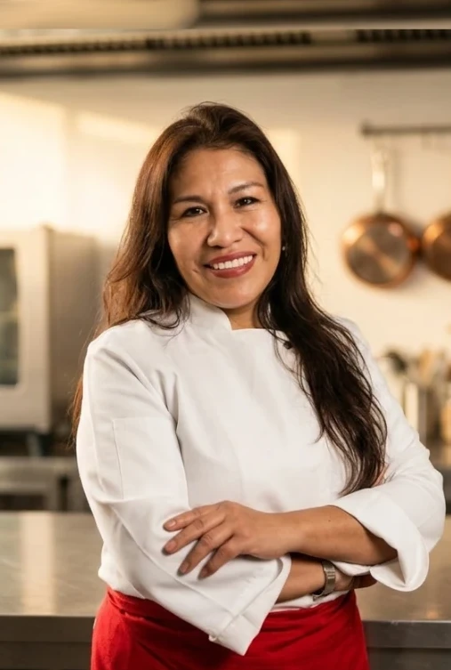 A smiling chef in a white jacket stands with folded arms in a professional kitchen.