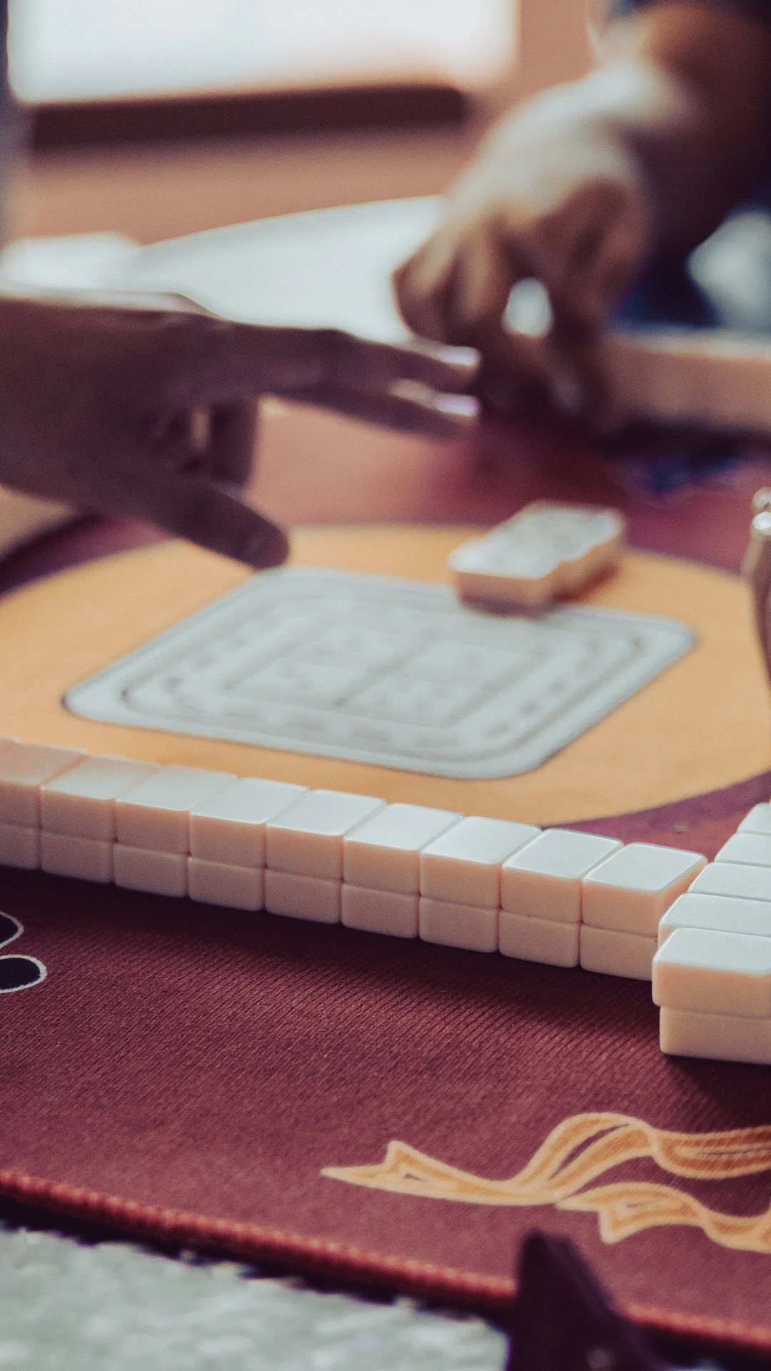 Hands of people playing Mahjong on a decorated, burgundy table.