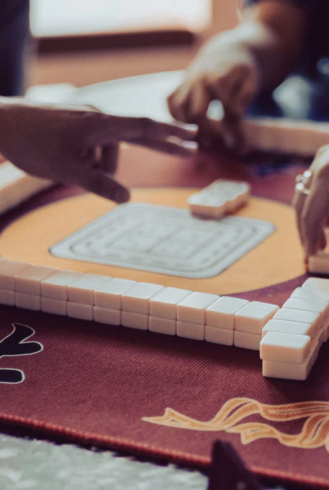 Hands of people playing Mahjong on a decorated, burgundy table.