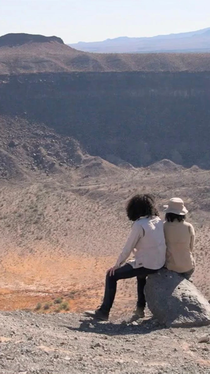Still from the movie: Two people sit on a rock overlooking a vast desert crater under a clear sky.