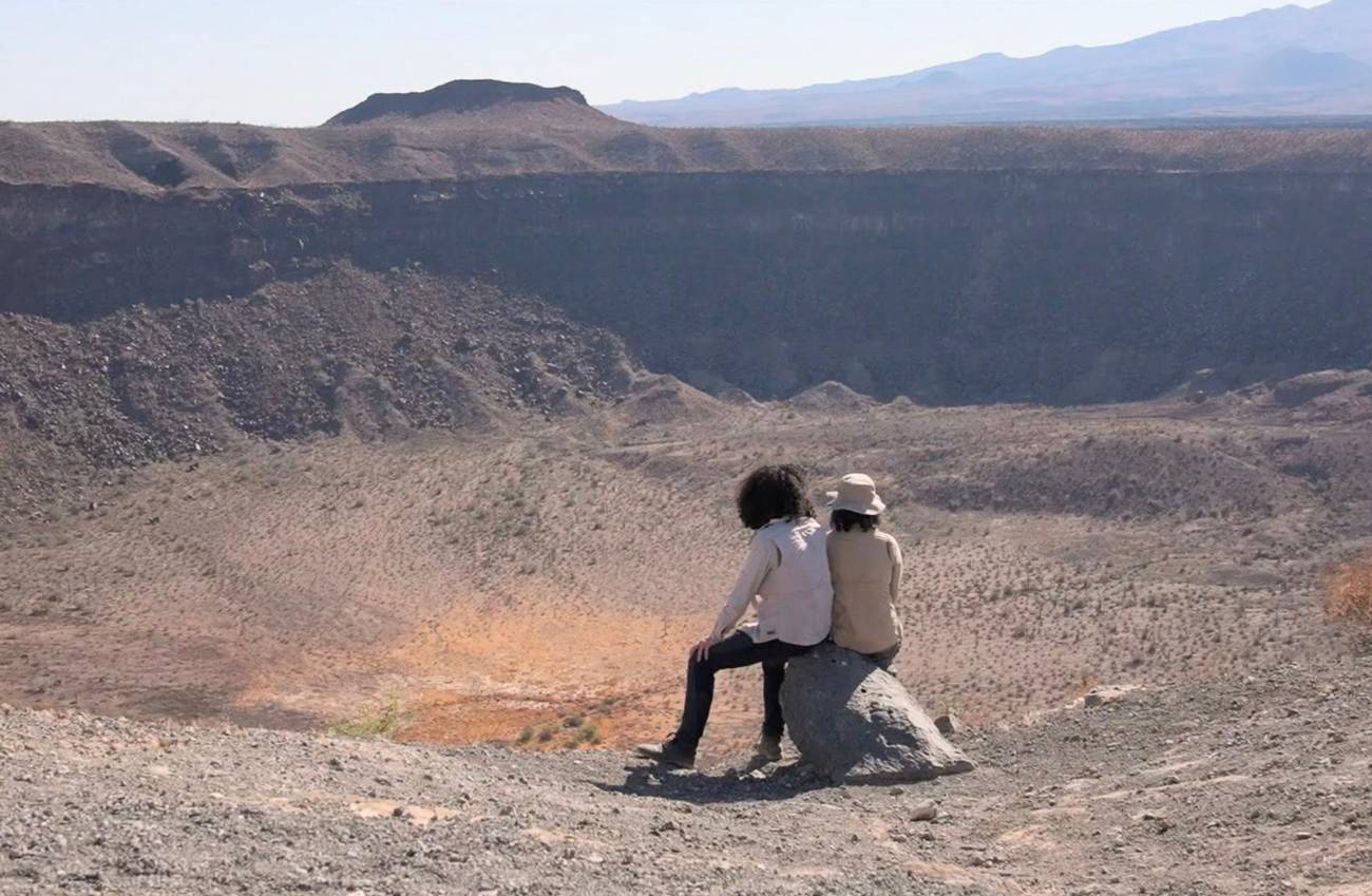 Still from the movie: Two people sit on a rock overlooking a vast desert crater under a clear sky.