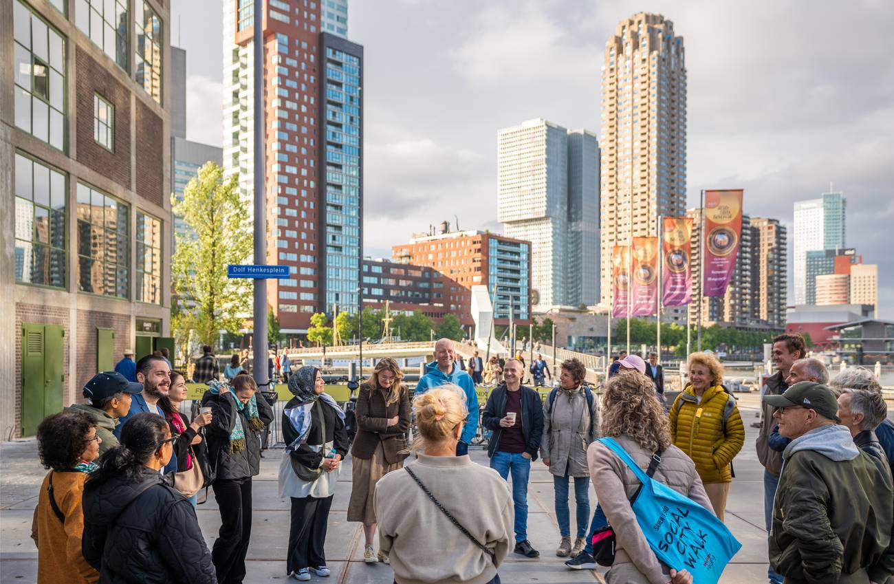 A diverse group of people stands in a circle on a city street, engaged and smiling. Tall buildings a
