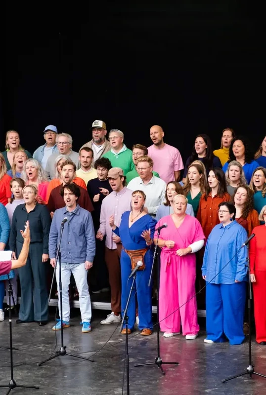 een koor met veel mensen gekleed in de kleuren van de regenboog zingt in een theater
