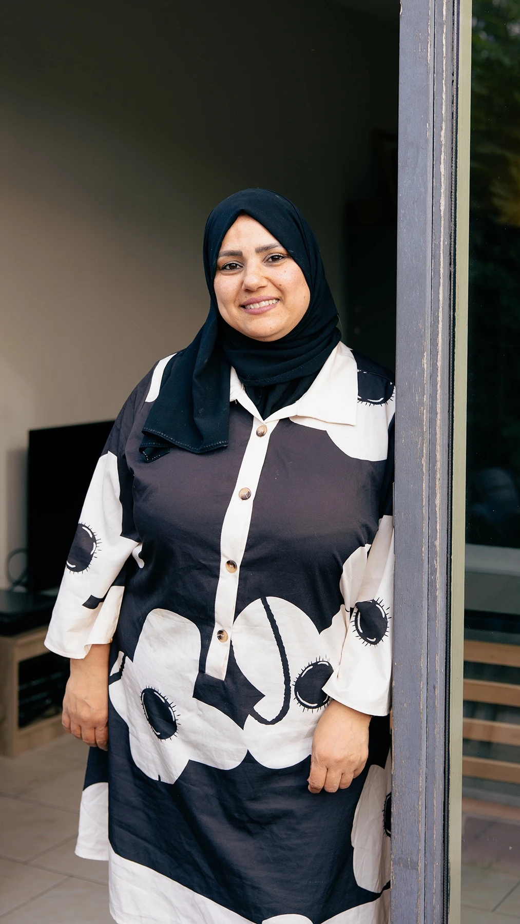 A woman wearing a black hijab and floral dress stands smiling in a doorway.