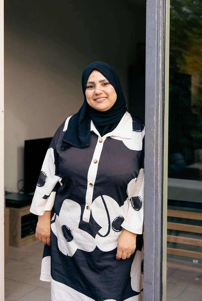 A woman wearing a black hijab and floral dress stands smiling in a doorway.