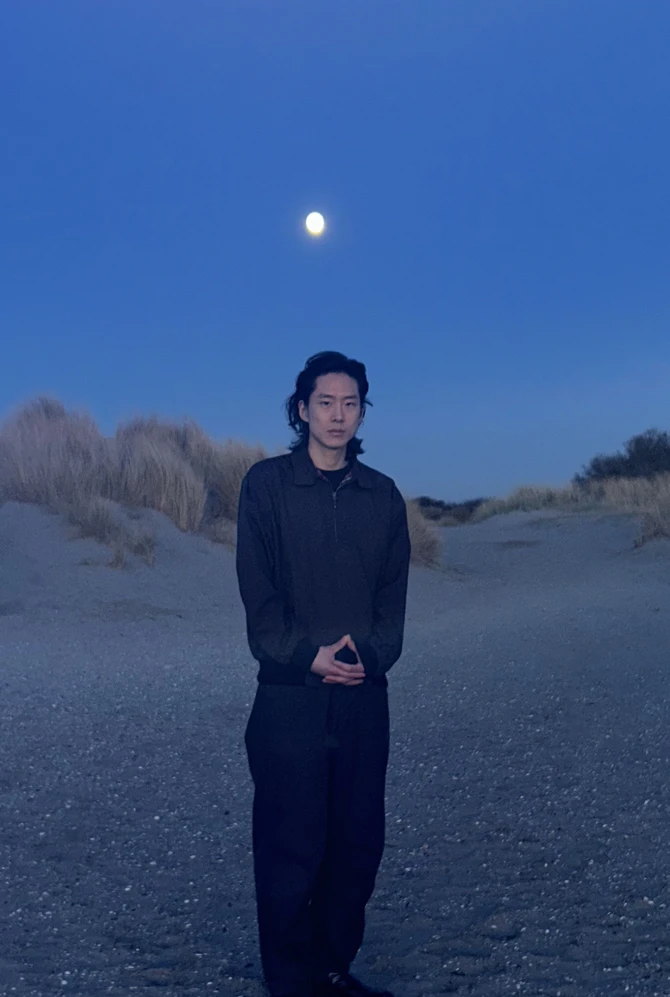 A person stands on a sandy beach at dusk under a clear blue sky, with the moon visible above. Grassy