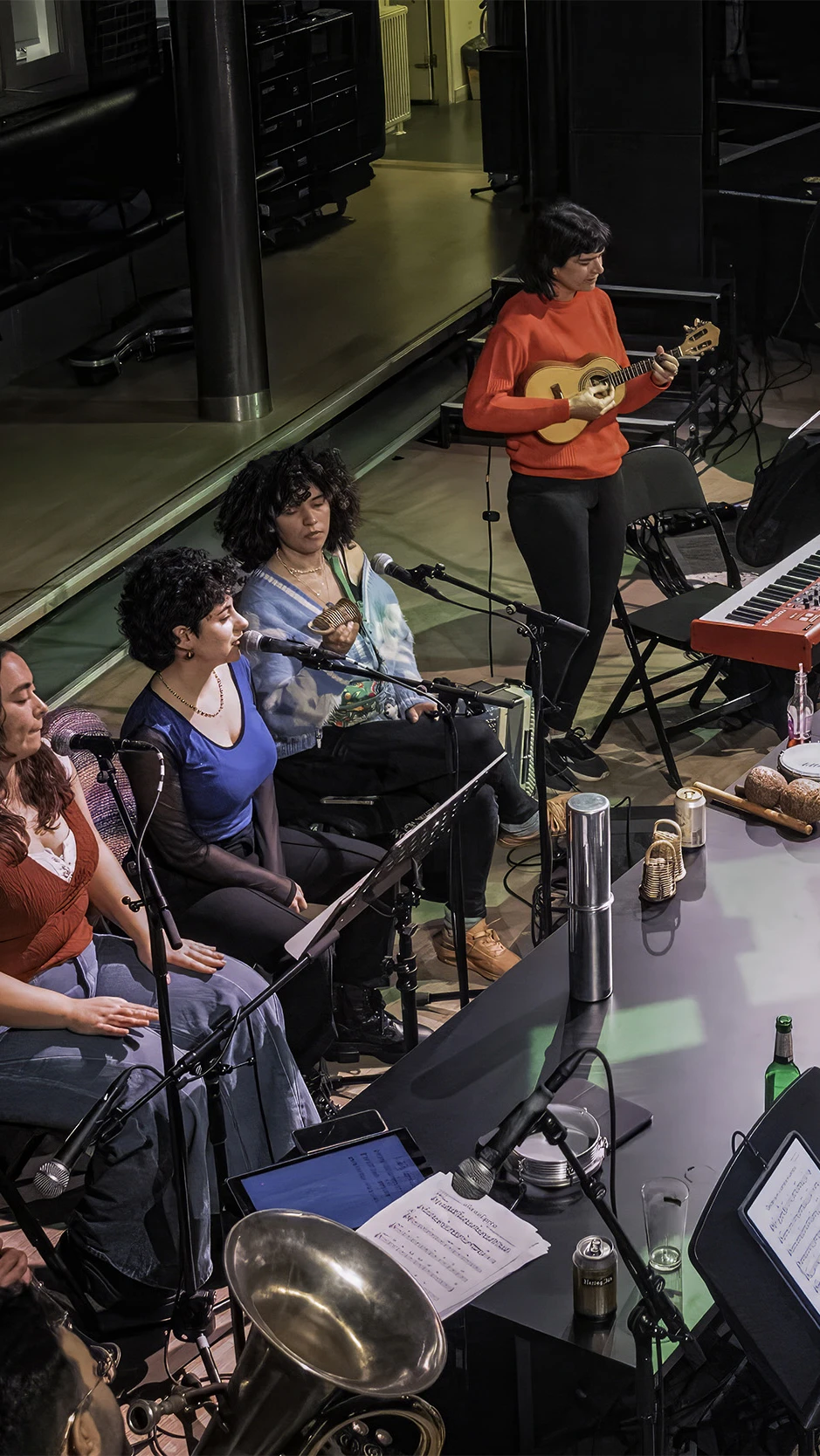 A diverse group of musicians rehearses around a long table with instruments like a keyboard, guitars