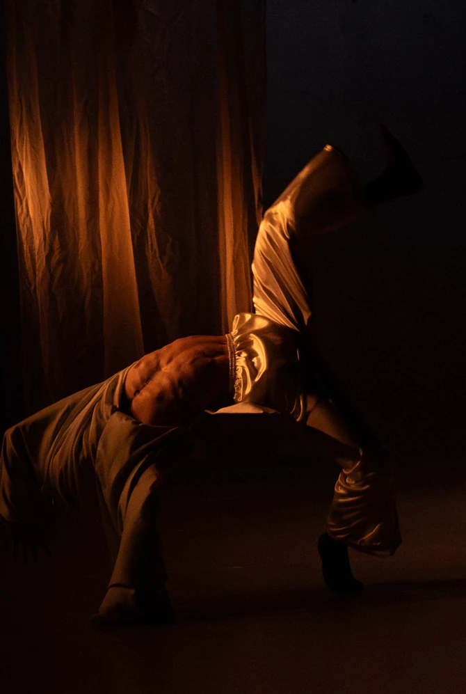 A dancer performs a dynamic move, in dramatic lighting. The scene is dimly lit with warm tones.