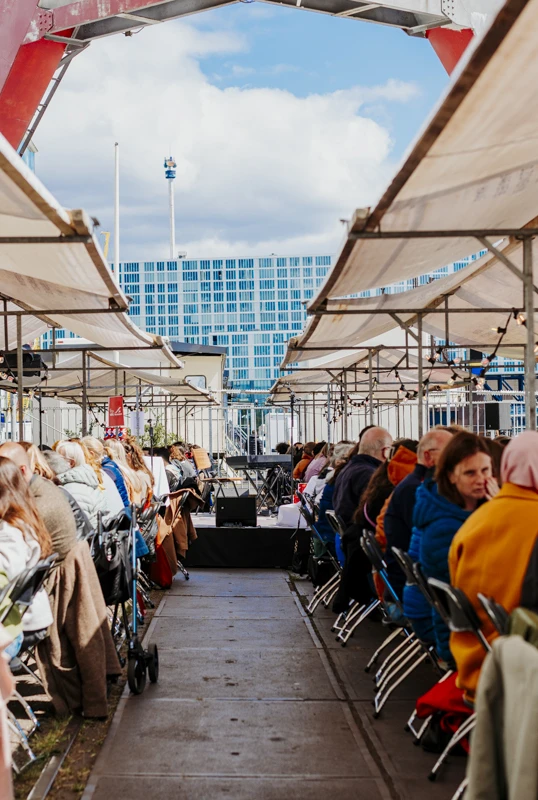 Outdoor dining scene with long tables under white canopies, filled with people in colorful attire.