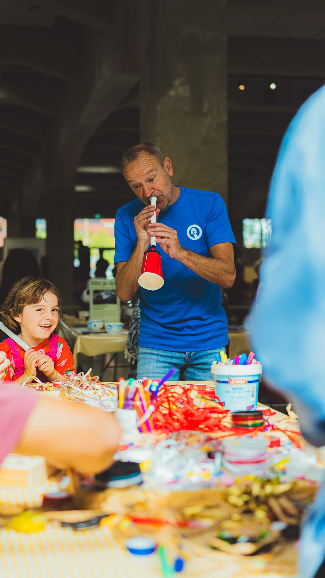 A man demonstrates a handmade musical instrument to children at a colourful craft table.