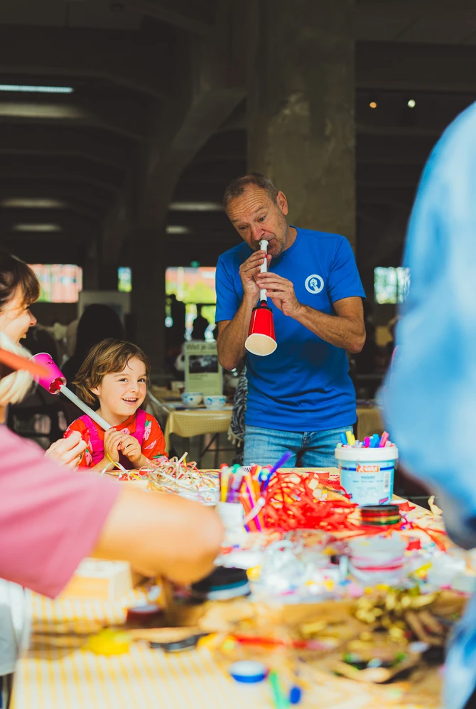 A man demonstrates a handmade musical instrument to children at a colourful craft table.
