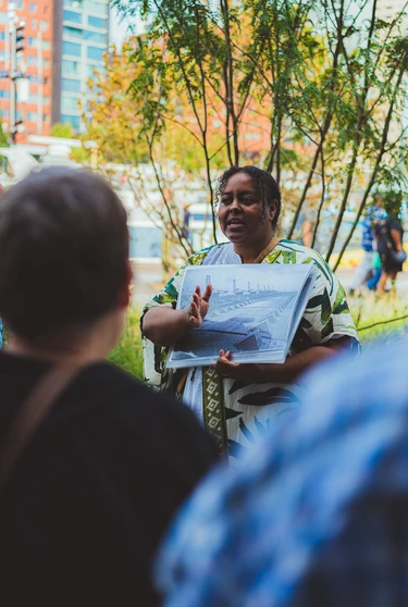 A woman speaks to a group outdoors while holding up a historical photograph of a harbour.