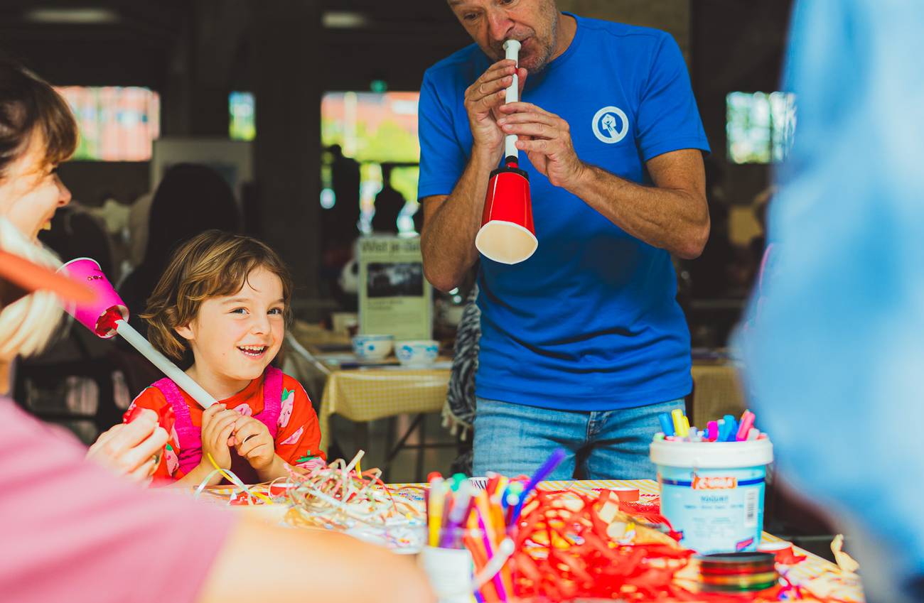 A man demonstrates a handmade musical instrument to children at a colourful craft table.