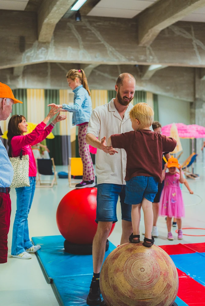 An adult helps a child balance on a large wooden ball during an indoor family activity.